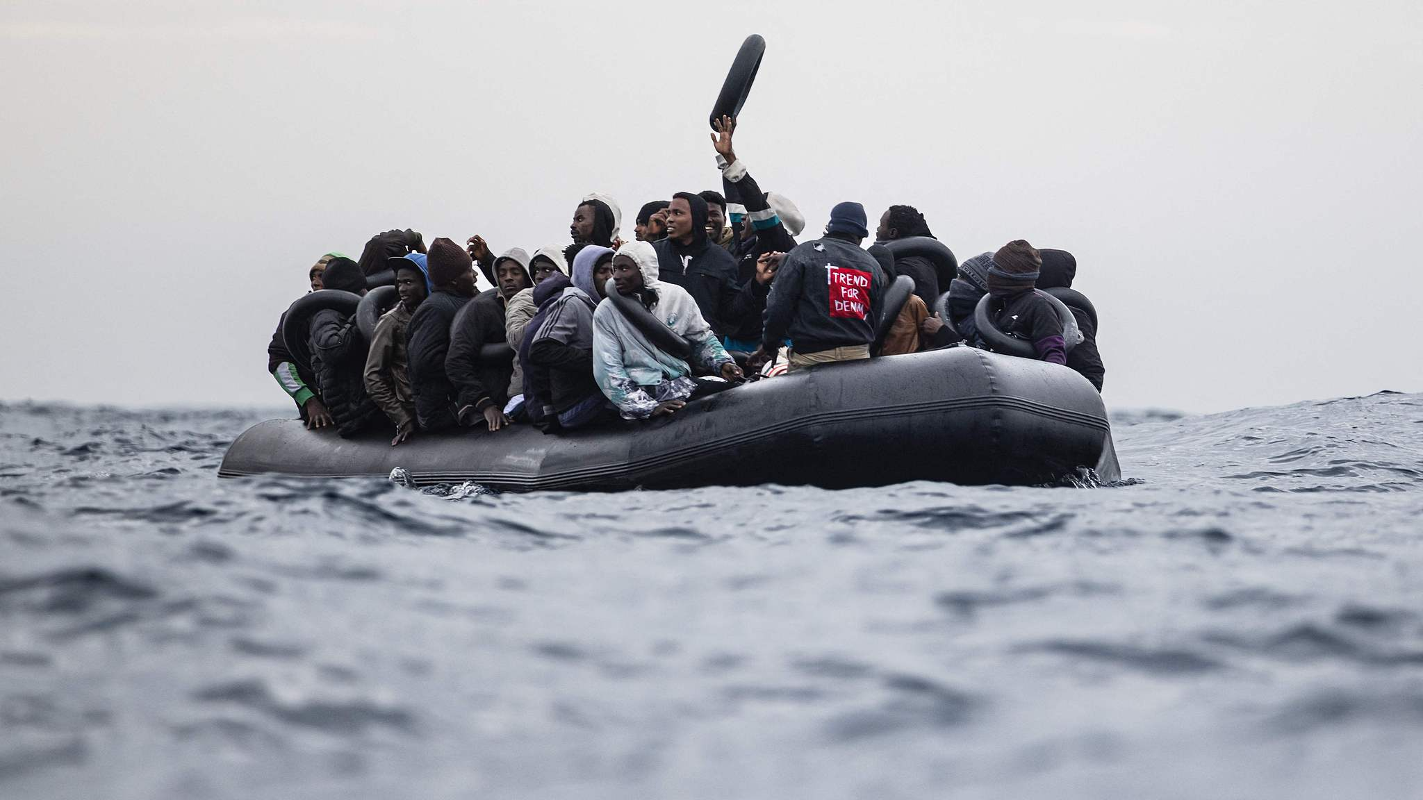 In the search and rescue zone near the Libyan coast in the Mediterranean Sea, migrants wave and gesture from their rubber boat, awaiting rescue by the crew of the 